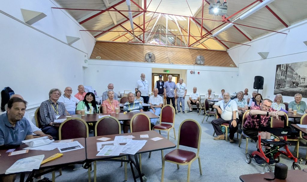 Group of people sat in a room gathered for a prostate cancer support group meeting. There is a few empty chairs and an empty table at the front of the picture.