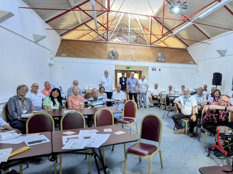 Group of people sat in a room gathered for a prostate cancer support group meeting. There is a few empty chairs and an empty table at the front of the picture.