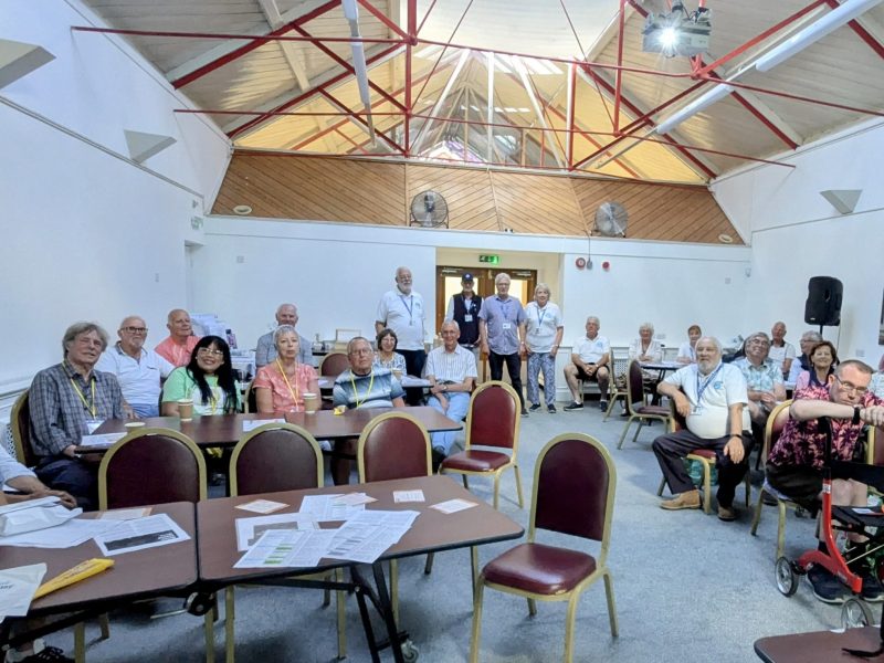 Group of people sat in a room gathered for a prostate cancer support group meeting. There is a few empty chairs and an empty table at the front of the picture.
