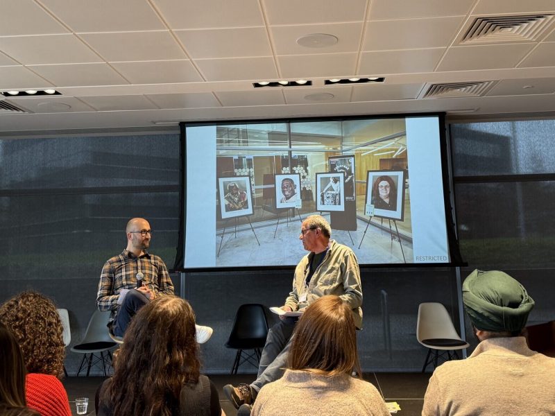Two men sat on chairs on a stage in front of a screen, which shows images of a photography exhibition.