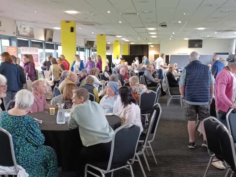 Big group of people sat around tables chatting to each other, in a community setting.