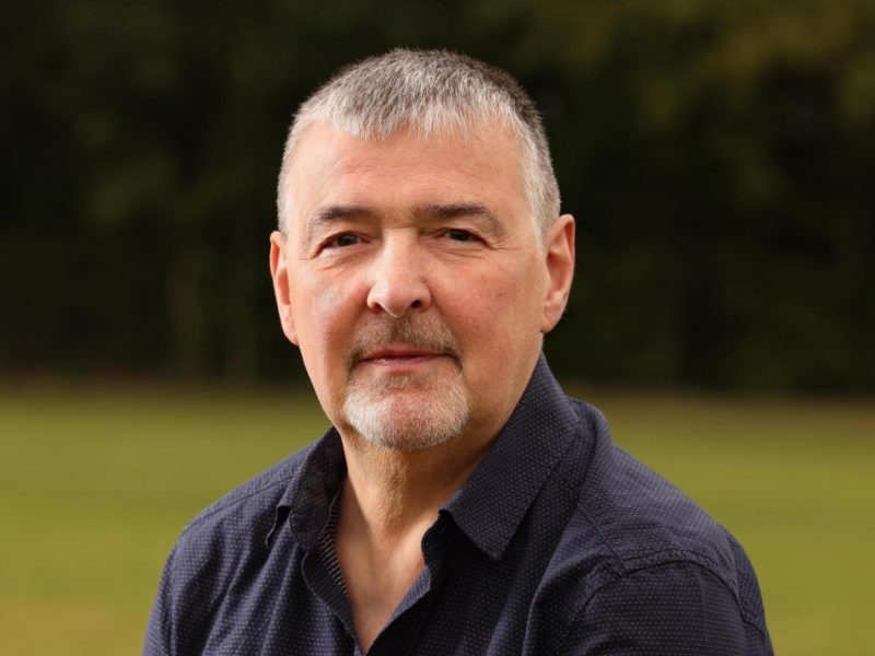 Tony Collier, vice-chair of Tackle Prostate Cancer. Who is a white middle -aged man, sat looking into the camera. He is wearing a navy blue shirt and is sat in a field.